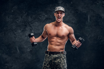 Fototapeta na wymiar Attractive shirtless man in cap is doing exercises with dumbbells and screaming.