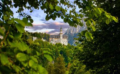 Fairy-tale Neuschwanstein Castle in Bavaria, Germany. View through green leaf of tree at famous vintage landmark. Picturesque evening sunset landscape.