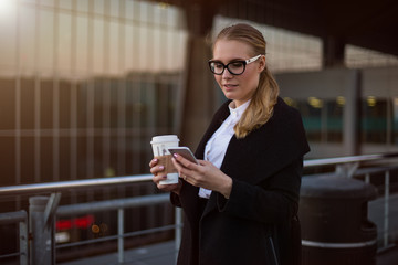 Smiling successful businesswoman online banking via mobile phone while standing with take away coffee outside company after work day. Female skilled broker sending sms via cell telephone gadget