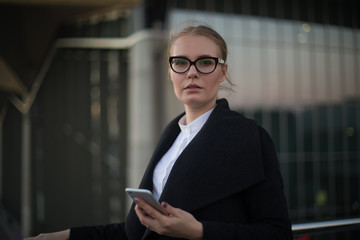 Portrait of a serious confident woman professional banker in fashionable spectacles and autumn coat holding in hand mobile phone and looking in camera while standing against modern building