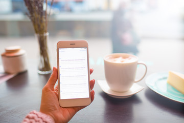 Close up female blogger reading information on web site via mobile phone with blurred screen while relaxing in restaurant with cup of coffee. Woman student searching information via cell telephone