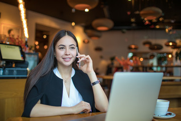 Happy smiling woman skilled leadership talking with partner via cell telephone while sitting with pc laptop computer in restaurant during break at job. Joyful female manager phoning via smartphone