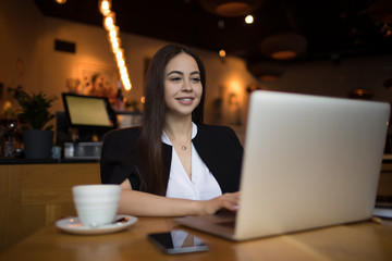 Smiling woman college student with good mood having online webinar via laptop computer during leisure time in restaurant. Joyful female marketing coordinator checking e-mail on netbook