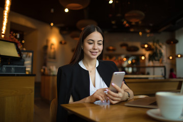 Charming woman in luxury wear checks the status of the account on mobile phone while resting in coffee shop during free time. Smiling pretty female manager online chatting on cellphone