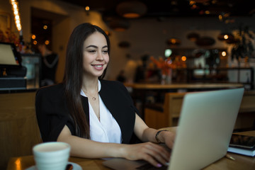 Happy smiling woman in fashionable clothes having online training course on laptop computer while sitting in coffee shop during work break in company. Joyful female blogger typing lifestyle article