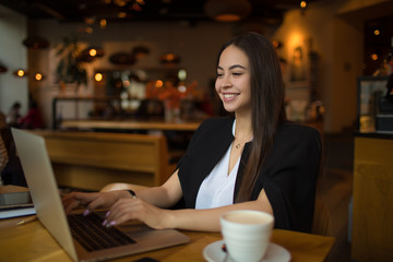 Young smiling woman clever university student in official wear having online education via portable pc laptop computer while resting in restaurant. Cheerful female magazine editor typing on netbook