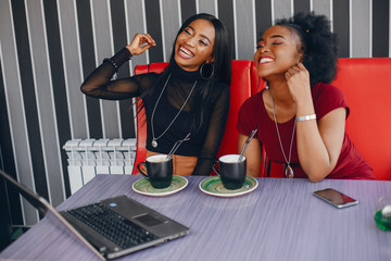 two beautiful and stylish young, dark girls sitting in a restaurant at the table, chatting and using a laptop and drinking a coffee