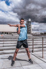 Fototapeta premium Athlete man doing shoulder muscle stretch before training. In the summer in the city, preparation for fitness workout, sportswear shorts t-shirt and sneakers. Step cloud background.