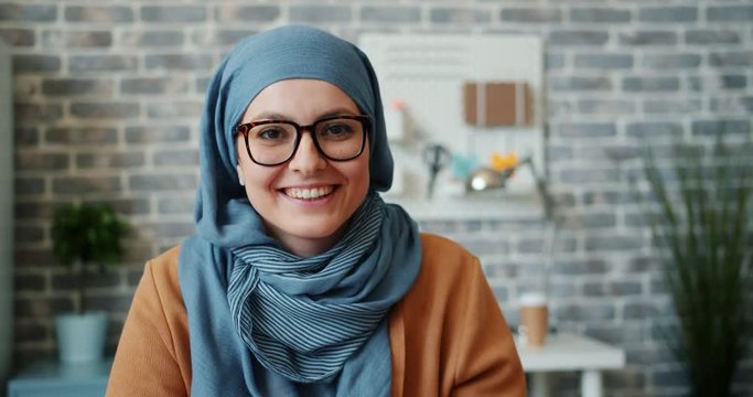Slow motion of pretty girl in hijab and glasses smiling looking at camera in office alone. Modern young people, creative workplace and business ladies concept.