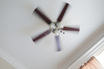 Ceiling fan with a ceiling lamp turned on, working with blurred blades in a living room, ceiling with moldings, curtain with shades and rods.