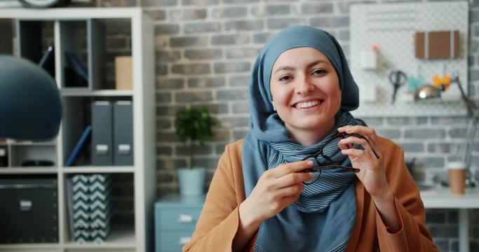 Portrait of attractive young Muslim businesswoman in hijab holding glasses sitting in office alone looking at camera with happy face. People and business concept.