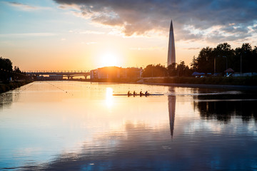 Boat regatta/rowing team silhouette on the tranquil lake at sunset, cityscape on background. 