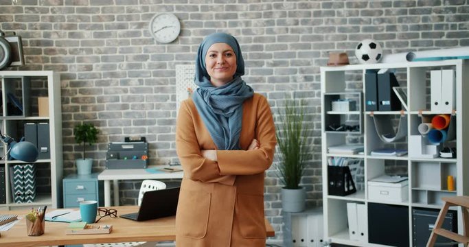 Slow motion portrait of successful Muslim businesswoman smiling in office standing alone with arms crossed looking at camera. People, work and success concept.