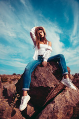 Beautiful girl in a white shirt, blue jeans and cowboy hat posing on the rock. Outdoor shot. Summer vacation concept.