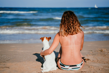 young woman and her cute small jack russell terrier sitting at the beach enjoying and relaxing. Summer and holidays concept