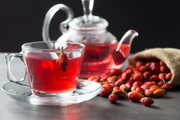 Dried rose hip glass cup of herbal tea medicinal plants with rosehip fruits in burlap sack and glass teapot on black rustic table. Dogrose winter tea background