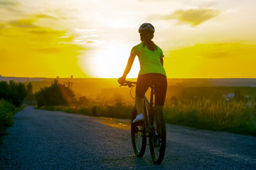 Naklejka na ściany i meble Beautiful girl cyclist riding a bike on the road towards the sunset.