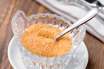 crystal bowl with brown cane  sugar,  spoon and white saucer macro (shallow focus) on wooden table 