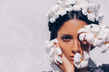 close up of beautiful young woman wearing wreath and holding frangipani flowers near face at white...