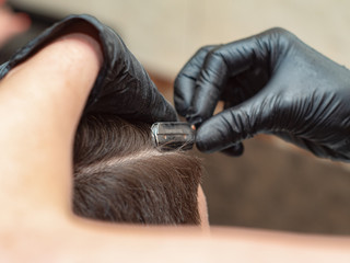 Hairdressing stylist cutting hair with straight razor, close up view. Females hands in black rubber gloves with open razor. Photographed in barbershop. Selective soft focus. Blurred background