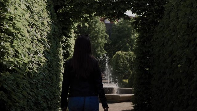 Long-hair girl goes to the fountain along the alley in the maze of green bushes in a garden. Back view. Woman resting outdoors on weekends. Beautiful landscaping. Garden design