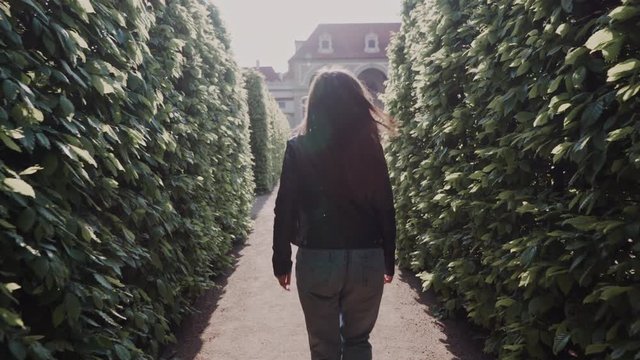 Beautiful smiling girl walking in park labyrinth in evening sunshine. She turns around and smiles at the camera. Back view