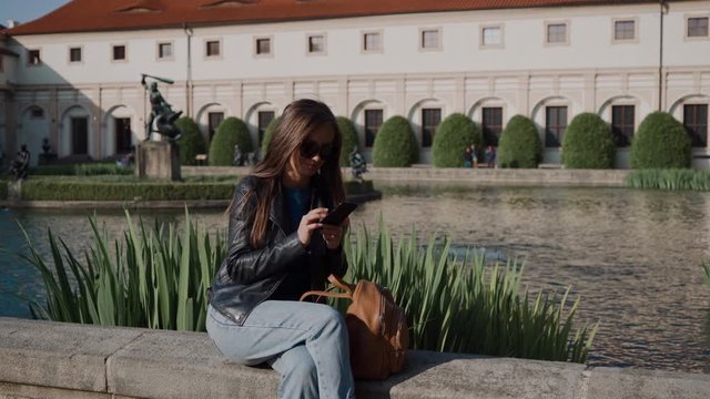 Young attractive girl using smartphone while sitting in beautiful park by the pond. There are Wallenstein Palace and the pond with fish and sculptures on blured background. Beautiful landscaping