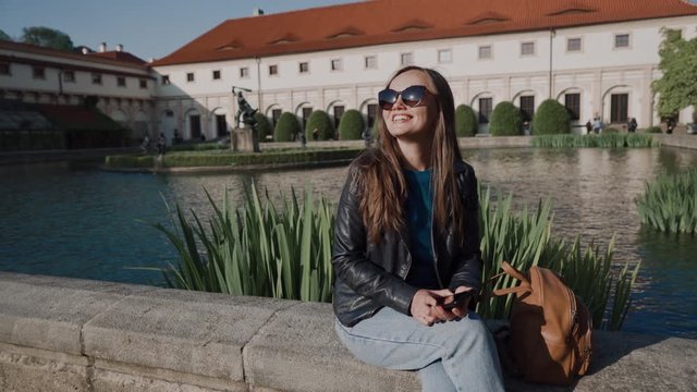 Portrait of beautiful smiling woman with sunglasses with a phone in her hands sitting near the pond in the park in evening sunshine. There are Wallenstein Palace and the pond with fish and sculptures