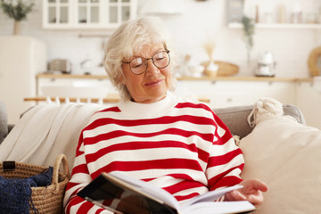 Beautiful gray haired senior European woman in stylish round eyewear enjoying reading novel,...