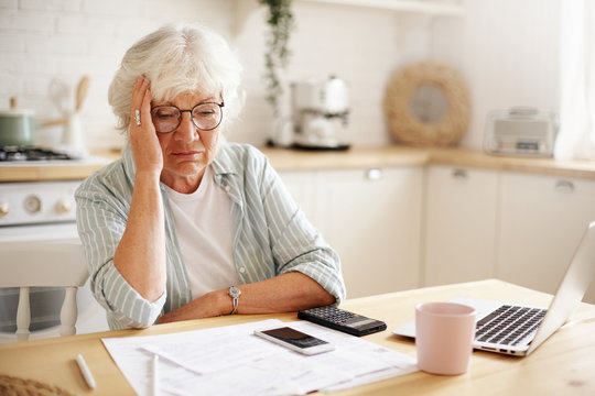 Sad frustrated senior woman pensioner having depressed look, holding hand on her face, calculating family budget, sitting at kitchen counter with laptop, papers, coffee, calculator and cell phone