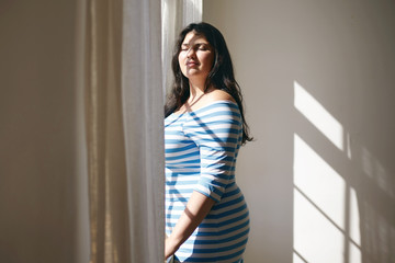 Fototapeta na wymiar Portrait of charming sensual Latin plump female in striped dress closing eyes in enjoyment, feeling bright warm sunshine on her face, standing by window in bedroom. People and lifestyle concept