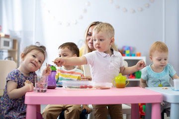 Kids have a dinner in kindergarten. Little toddlers boys and girls from the group of children sitting at the table with lunch and eat appetizing. Children with caregiver in day care centre