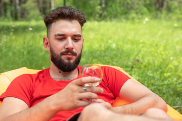 Naklejka na ściany i meble young hipster bearded guy, man lying or relaxing on an inflatable sofa, holds in his hand a glass of wine. .against the background of green landscape