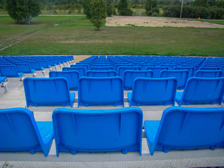 Lot of plastic seats. Back view. Field with green trees and grass.
