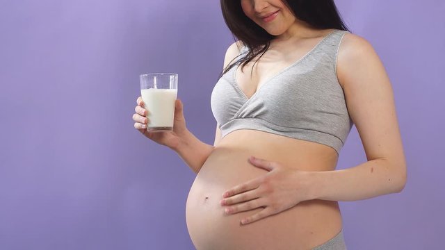 Pregnant slender woman in lingerie with big belly holds a glass of milk isolated over blue wall.