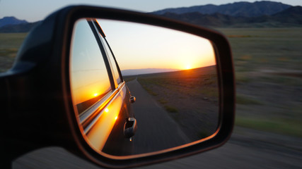 View in the side mirror of the car. Orange dawn beyond the hills. The car goes at speed. Visible green fields, grass, grasslands. Black color of the car.