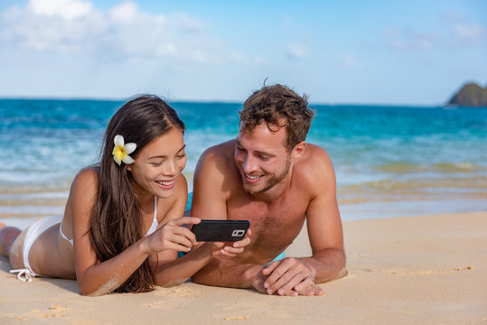 Beach Holiday Couple Sunbathing Relaxing Having Fun Using Phone App 5g Data. Young Tourists Friends Technology Lifestyle. Happy Multiracial Asian Caucasian Biracial Family Enjoying Summer Lifestyle.