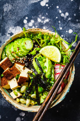Poke bowl with avocado, black rice, smoked tofu, vegetables, sprouts, dark background.