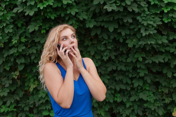 Beautiful young girl in a blue dress talking on the phone. Against the background of wild grapes, summer day. Copy space.