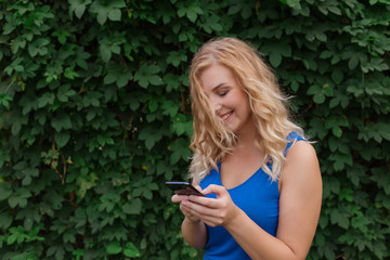 Beautiful young girl in a blue dress typing a message on the phone. Against the background of wild grapes, summer day. Copy space.