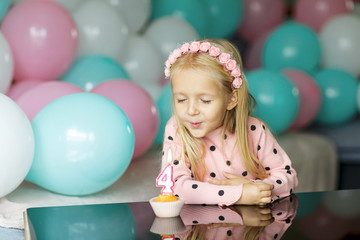 Fototapeta na wymiar Indoor shot of pretty joyful little girl with blonde hair blowing out the candle, celebrate 4 years old birthday, wear fashionable dress, have excited expressions. Happy childhood concept