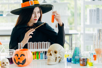 Attractive beautiful Asian woman dressed as a witch using magic wand casting spell to potion in test tube at modern laboratory, Halloween holidays.
