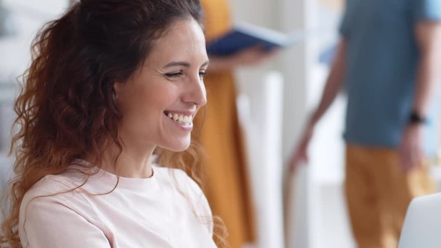 Close up of Caucasian woman with curly hair and headphones in her ears sitting at laptop, talking via internet with interlocutor while her colleagues communicating on background