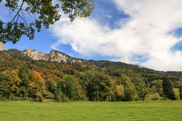 View of the mountains in early autumn. In the foreground is a lush green meadow. Trees have already got something of the red and the yellow color. The sky is full of white clouds.