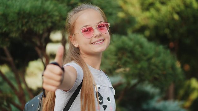Happy young schoolgirl in pink sunglasses showing thumb up, the girl smiling portrait, face outdoor in park. Slow-motion 4k video