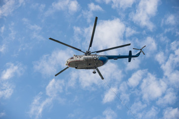 A white and blue helicopter flying in the sky - the cable hangs out of the helicopter
