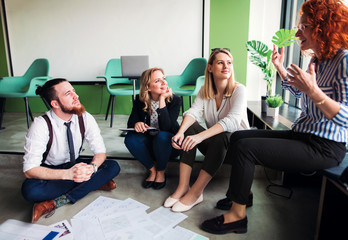 A group of young business people sitting on the floor in an office, talking.