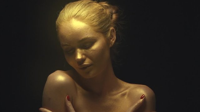 Beautiful sexy girl with golden skin. Fashion model woman in golden bright sparkles posing in studio. Creative metallic gold makeup. Slow motion 4K UHD video footage. 3840X2160
