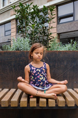 Little girl make yoga in modern city.  Sitting in lotus position on bench. Harmony concentration.