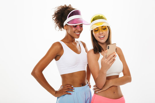 Portrait of two cute multinational women wearing visor hats smiling and using cellphone